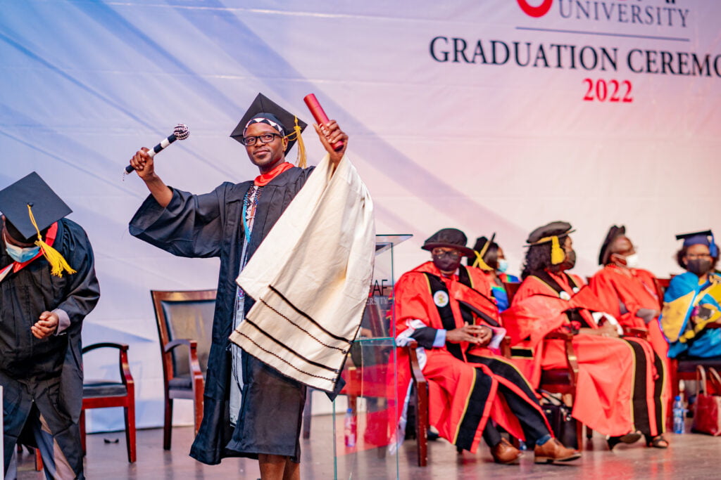 Graduate on a stage wearing graduation gown and cap holding a diploma