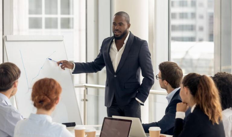 Businessman in dark suit presenting at whiteboard to a group of professionals in a bright modern meeting room.